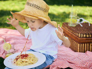 Boy in straw hat eating spaghetti outdoors on picnic blanket. Concept of natural parenting,...