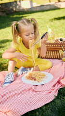 Girl in yellow outfit eating spaghetti and cutting it with scissors during outdoor picnic. Concept...