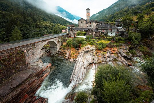 view of Lavertezzo village with a stone bridge over Verzasca river and lush foggy green mountains - Powered by Adobe