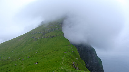 Vibrant green cliffs rise dramatically from the ocean beneath a cover of rolling clouds in the Faroe Islands. The serene atmosphere invites exploration, Kallur Lighthouse, Kalsoy Island
