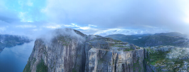 Experience the breathtaking panorama from Preikestolen Cliff, where towering cliffs meet the serene waters of Lysefjord. Clouds drift by, adding a mystical touch to this stunning Norwegian landscape.