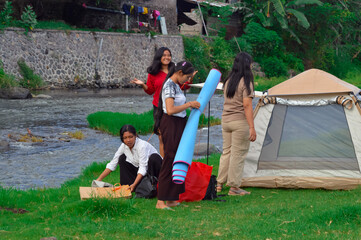 Four teenage girls prepare their riverside campsite, one unrolling a mat while another crouches to...