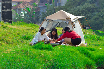 Girls at riverside tent camping place sausage skewers one by one onto the hot grill, while others...