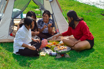 By the riverside tent, teenage girls watch sausage skewers cook on the portable stove while...