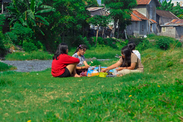 Four teenage girls sit on the riverside meadow, grilling skewered sausages on a portable stove...
