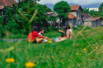 Four teenage girls sit on the riverside grass, grilling skewered sausages on a portable stove with...