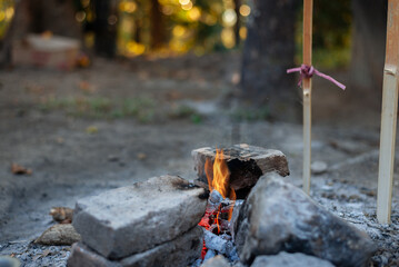 Fire burns between stacked stones, with glowing embers and a visible flame, set in a natural environment with trees and soft sunlight in the background.