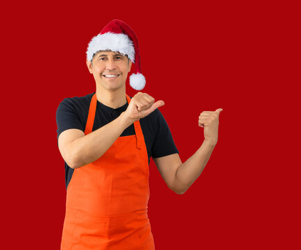 Studio shot of a hispanic barista with red apron standing over isolated red background smiling and looking at the camera pointing with two hands and fingers to the side while wearing a Christmas hat