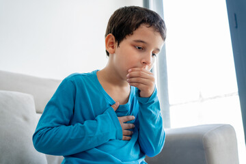 Little boy sitting on sofa at home and holding hand on chest. Male having asthma attack, difficulties with breathe, feeling severe pain or dyspnea