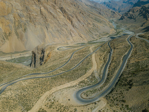 Aerial view of a winding road snaking through the arid, mountainous terrain, contrasting with the pale earth tones and sharp rock formations, Kaza, Himachal Pradesh, India.