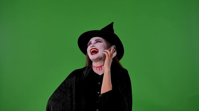 A witch with vampire makeup, in black cloak and hat, talks on her smartphone and laughing