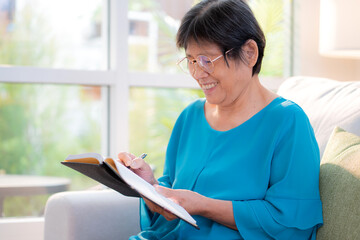 Asian senior woman writing book sitting on sofa with relax in living room at home, elderly woman planning and strategy writing diary with cozy, hobby and recreation in weekend, lifestyles concept.