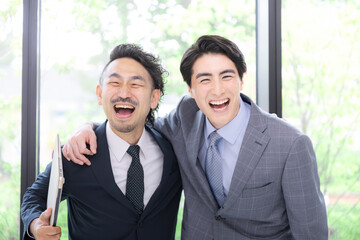 新緑の窓が綺麗なオフィスでビジネスマン肩を組む　達成や成功のイメージカメラ目線