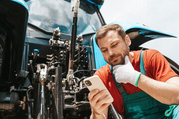 Modern farmer using digital technology in agriculture. Man using smartphone near tractor.