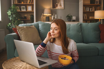 A woman sits on a couch with a relaxed expression, enjoying a bowl of snacks as she focuses on her...