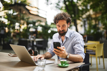 A man with curly hair is focused on his smartphone while working on a laptop at a lively outdoor cafe, surrounded by greenery and modern seating arrangements.