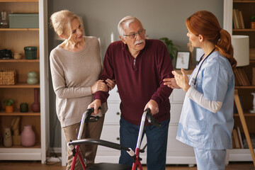 In a bright living room, a home health aide assists an elderly man using a walker. His wife supports his arm as the caregiver provides instructions or encouragement.