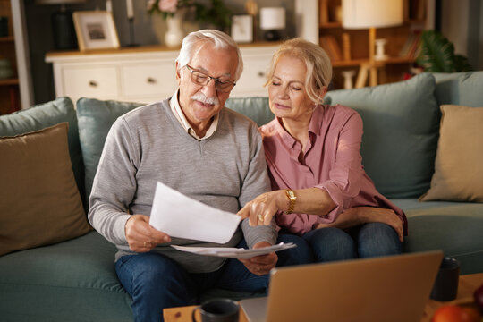 An elderly couple, a man and woman, sit on a blue sofa inside a home, reviewing papers related to financial planning, insurance, and retirement near a laptop.