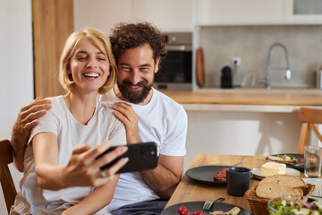 Smiling couple captures a fun selfie together during breakfast. They share a casual moment while seated at a table surrounded by food in a warm kitchen.