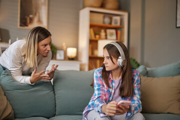 In a cozy living room, a teenager wearing headphones sits on a couch looking away while a parent...