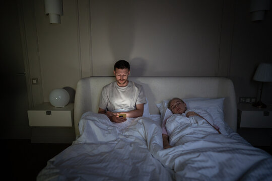 Stressed man engrossed in smartphone at night, disturbing wife sleep in bed. Male struggle with insomnia, phone digital addiction, endless scrolling through social media, sleep disruption, nomophobia