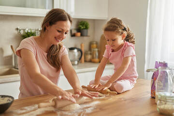 Mother and daughter work together at a wooden table, rolling dough and having fun baking. Flour is...