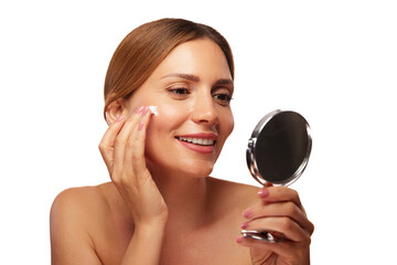 A woman smiles while applying skin care cream to her face, using a handheld mirror to check her reflection. She enjoys her morning beauty routine in natural light. © Stockphotodirectors