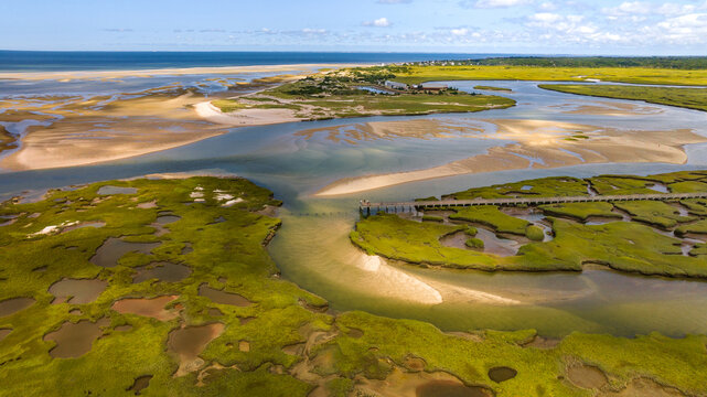 Aerial view of a long wooden pier over winding waterways that carve through vibrant green marshes and meet sandy Gray's Beach. Yarmouth Port, Cape Cod, Massachusetts, United States.