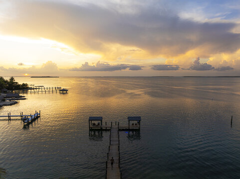 Aerial view of a long wooden pier stretching into the tranquil, sun-kissed waters beneath a sky ablaze with pastel hues, Key Largo, Florida, United States.