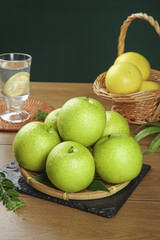 Fresh Green Apples on Bamboo Plate with Wicker Basket and Water Glass on Wooden Table