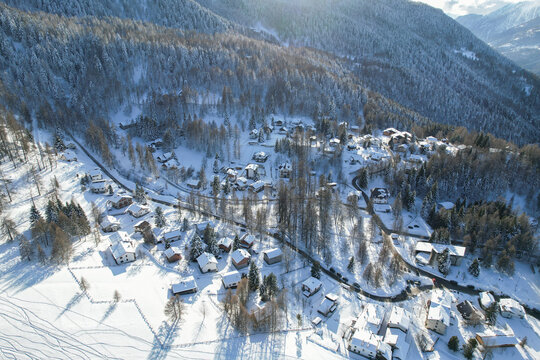 Aerial view of snow-blanketed houses nestled amidst frosted trees, a serene winter scene under the crisp morning light, Frais, Piemonte, Italy.