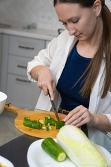 Concentrating on slicing fresh cucumber, a young woman prepares a healthy salad in her modern...
