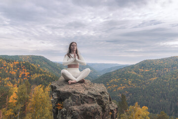 A white woman sits in the lotus position on a stone cliff, deep in meditation. Surrounded by...