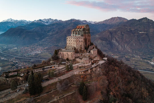 Aerial view of the Sacra di San Michele stands majestically atop a mountain, with the Alps providing a stunning backdrop under a pastel sky, Sacra di San Michele, Piemonte, Italy.