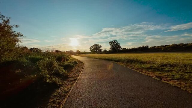 Cycling along open countryside trail at golden hour, POV. Riding bike through sunset lit field path, point of view. Moving forward on glowing road surrounded by grass, first person view