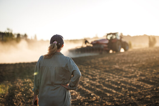 Farmer looking at tractor with seed drill sowing crop on field during sunset. Agriculture activity on farm