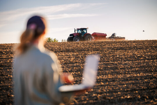 Tractor sowing crop on field while agronomist analyzing agricultural data on laptop. Smart farming in organic farm