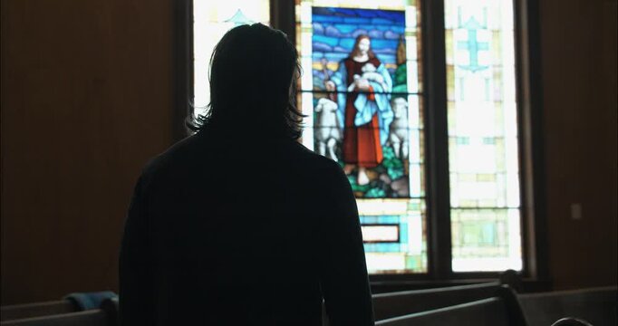 Back view of a man in dark clothing gazing at a stained-glass window, suggesting themes of redemption, purpose, and inner struggle.