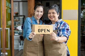 Two women wearing aprons smiling together at café entrance while holding an open sign,...