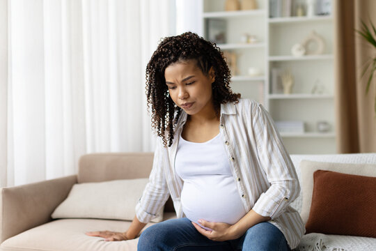 Concerned pregnant woman looks downward while holding her belly on the sofa. Theme of prenatal worry, uncertainty, and maternity challenges at home