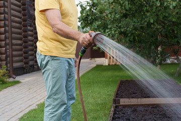 Senior man watering garden with hose in backyard. Organic farming and gardening concept,...