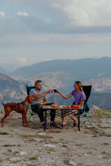 A young Caucasian couple enjoys a picnic on a mountain top. They toast with drinks while a brown dog sits nearby. Scenic mountains in the background.
