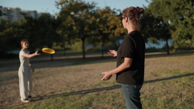 Female friends enjoying active leisure, throwing yellow frisbee across grassy park field surrounded by green trees under bright blue sky, showcasing playful summer recreation and bonding