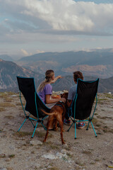 A young Caucasian woman and a young Caucasian man sit in camping chairs, enjoying a meal with a brown dog in the Caucasus mountains. Scenic mountain view in the background