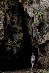 A young Caucasian woman with a backpack walks through a rocky cave surrounded by lush greenery. The scene captures the beauty of nature in the Caucasus region.