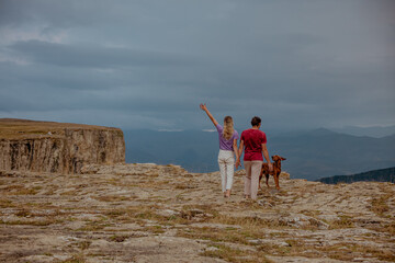 A young woman with long blond hair and a young man with short dark hair walk with a brown dog on a rocky landscape in the Caucasus mountains