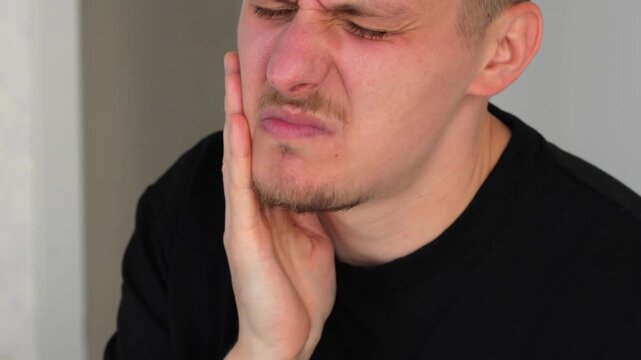 A patient with sore teeth stands in a room and holds his cheek with his hand.
Suffering from toothache in the mouth.
A man suffers from toothache