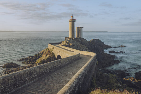 View of a stone causeway leading to the iconic Minou lighthouse perched on rugged rocks against the vast sea under a soft sky, Plouzan&Atilde;&copy;, France.