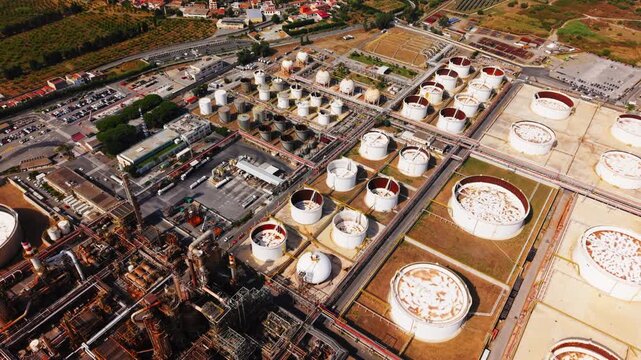 Drone in steep top-down angle capturing oil refinery tanks, pipelines, and surrounding roads in Sicily.