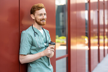 Ginger man doctor in scrubs drinking takeaway coffee near hospital wall